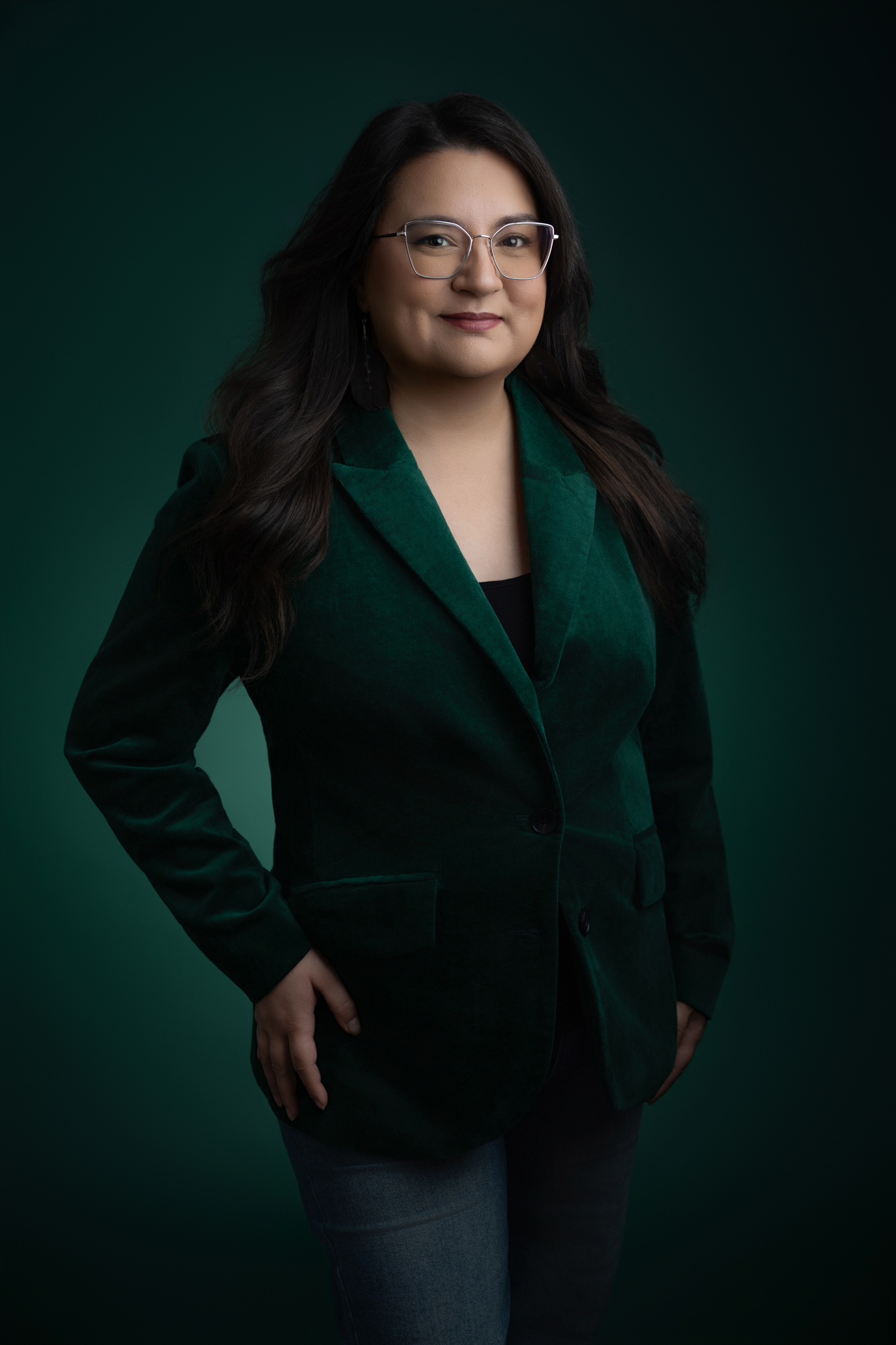 A woman with long dark hair and glasses wears a dark green blazer and jeans, standing against a dark green background—a perfect example of stylish headshot photography for Headshot Minis or quick mini sessions.