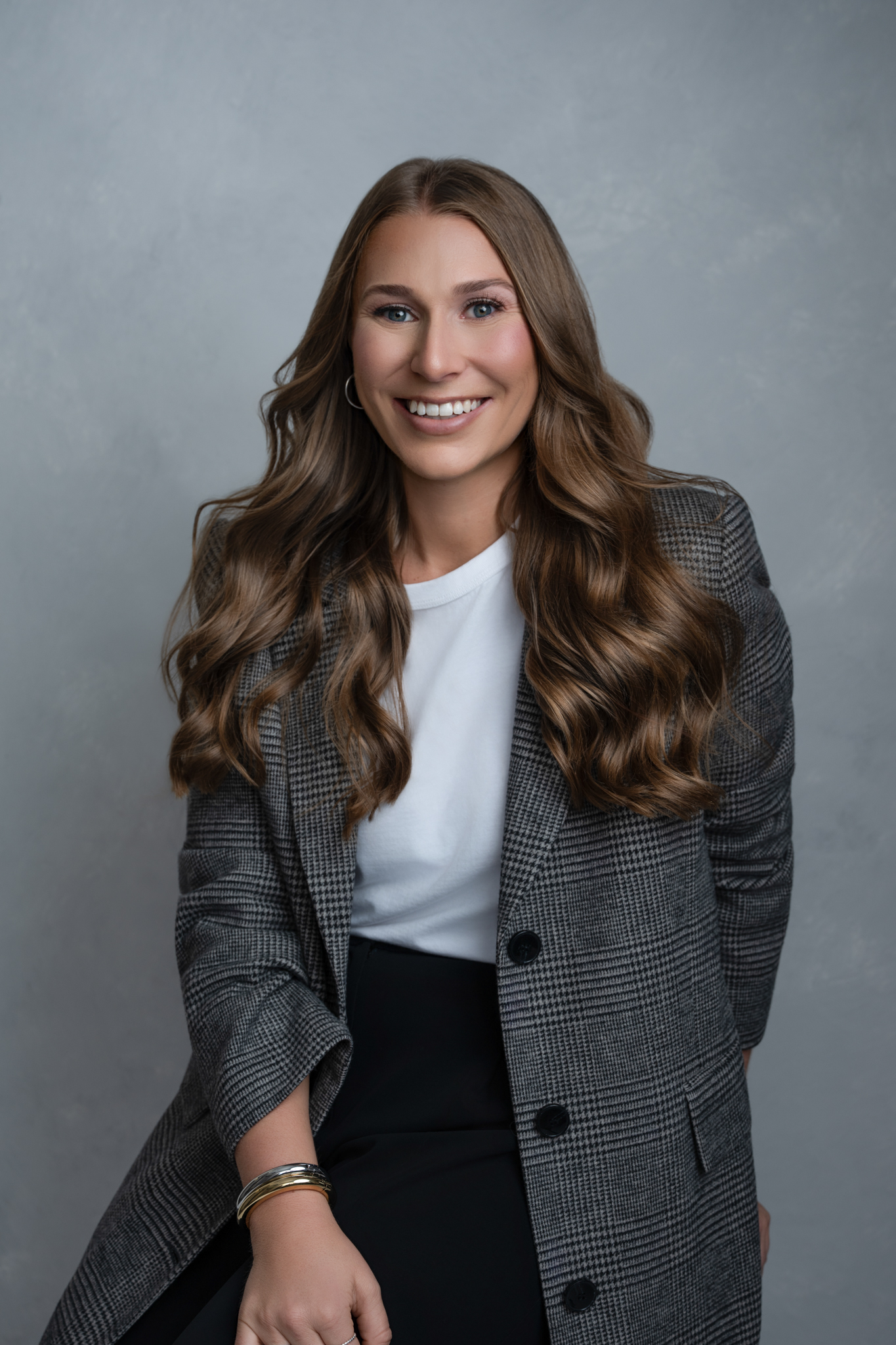 A woman with long wavy hair wearing a plaid blazer, white shirt, and black pants sits and smiles at the camera against a plain grey background, perfect for headshot minis.